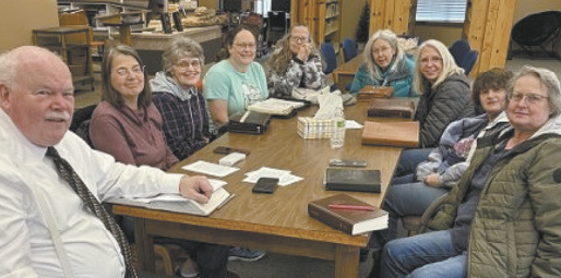 A group of people are sitting around a table, engaged in conversation. There are several books on the table, and a cup is placed in the center. The people are wearing ties, suggesting a formal or professional setting.

AI generated content